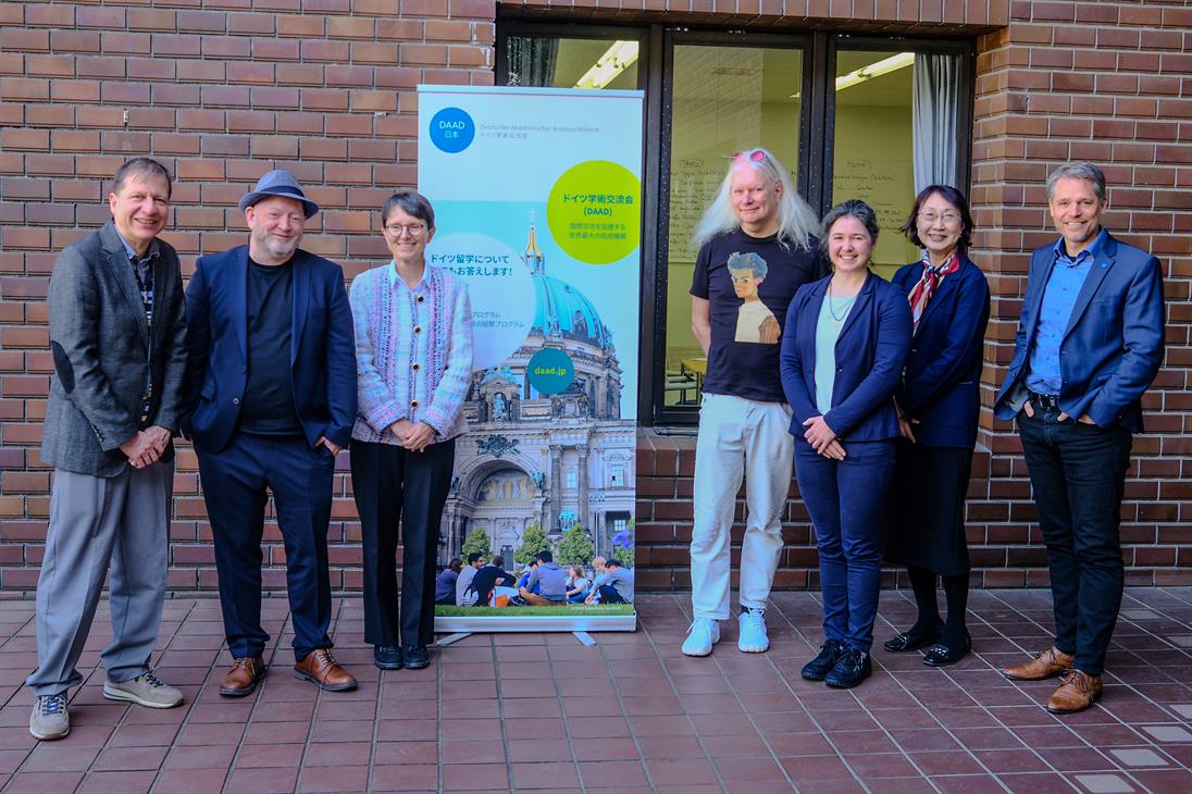 Eine Gruppe von sechs Personen steht vor einem Plakat, das Informationen über den Deutschen Akademischen Austauschdienst (DAAD) enthält. Das Plakat zeigt ein Bild eines Gebäudes in Deutschland mit dem Berliner Dom im Hintergrund und Menschen, die im Freien sitzen. Die Personen auf dem Foto sind in lockerer Kleidung gekleidet, darunter Jacken, Hosen und Mützen. Sie stehen vor einer Backsteinmauer und einem Fenster. Die Atmosphäre wirkt freundlich und gesellig, möglicherweise bei einer offiziellen oder akademischen Veranstaltung.
