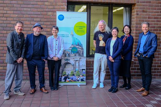 Eine Gruppe von sechs Personen steht vor einem Plakat, das Informationen über den Deutschen Akademischen Austauschdienst (DAAD) enthält. Das Plakat zeigt ein Bild eines Gebäudes in Deutschland mit dem Berliner Dom im Hintergrund und Menschen, die im Freien sitzen. Die Personen auf dem Foto sind in lockerer Kleidung gekleidet, darunter Jacken, Hosen und Mützen. Sie stehen vor einer Backsteinmauer und einem Fenster. Die Atmosphäre wirkt freundlich und gesellig, möglicherweise bei einer offiziellen oder akademischen Veranstaltung.